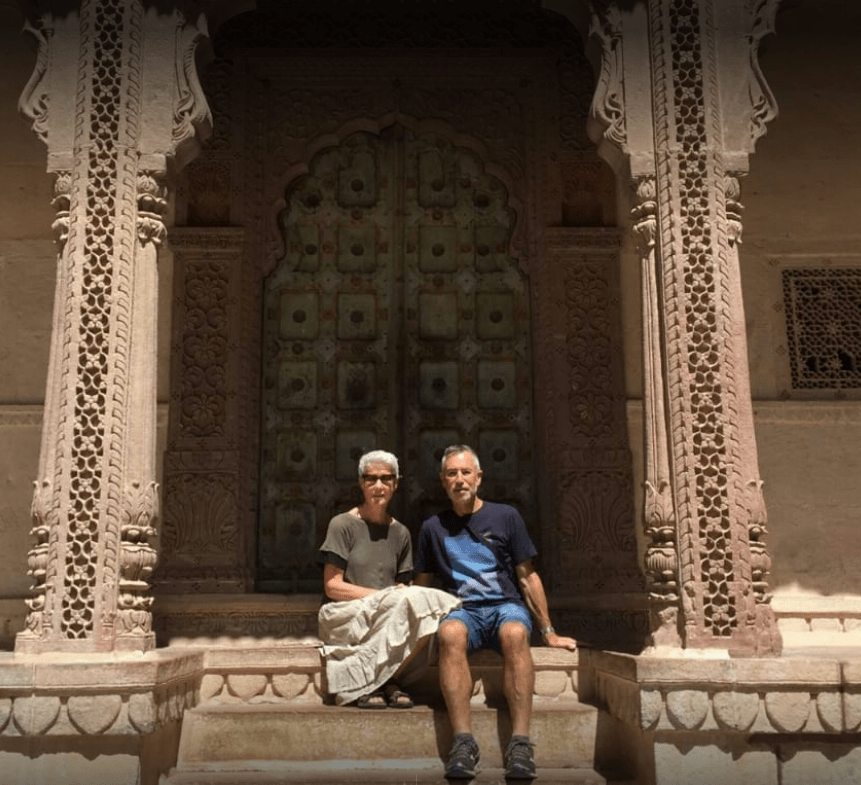 Two individuals seated on steps before an ornate building in India, showcasing intricate architectural details.